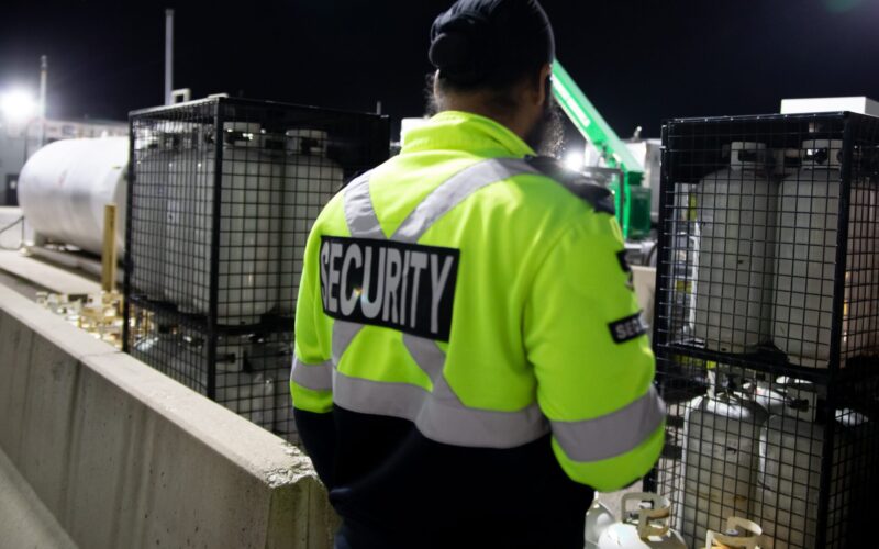 security guard in a neon yellow, silver, and black jacket that says “security” on the back stands overlooking an industrial property with tanks locked in black cages