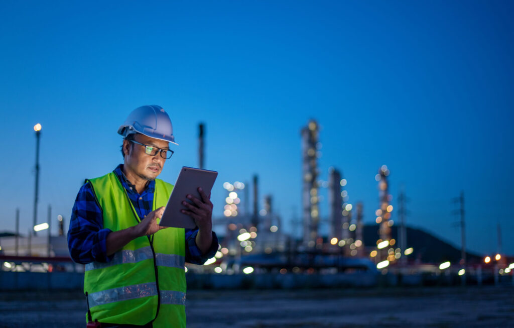 A worker in a safety vest and hard hat monitors security on a mobile device with the silhouette of a chemical plant in the background 