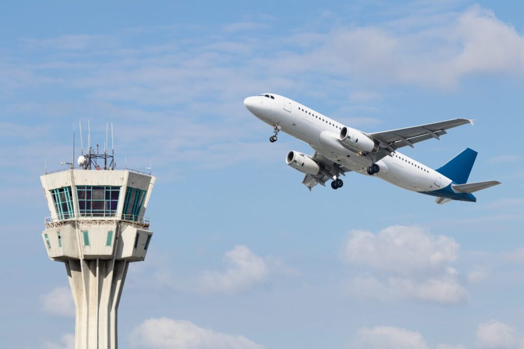 A plane takes off from the runway with the control tower behind and blue sky in the background.