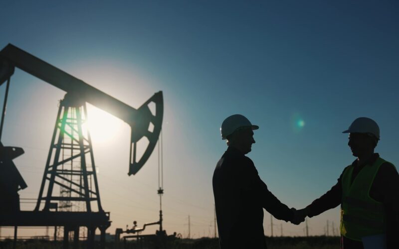 A twilight sky frames the silhouette of an oil rig and two businessmen shaking hands.