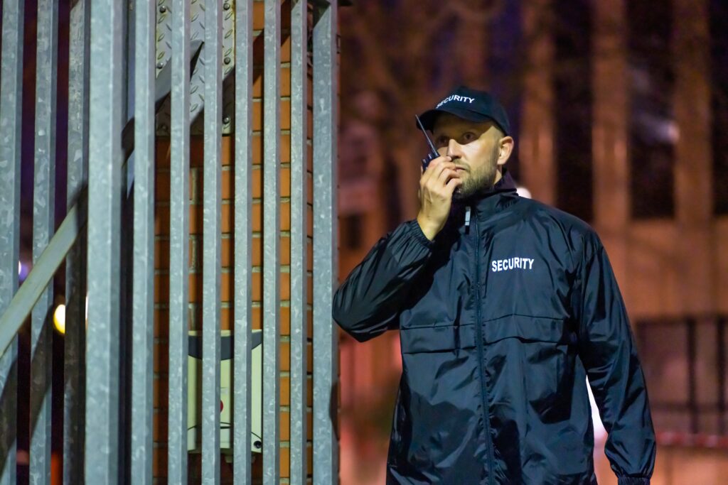 A security guard in a navy jacket and hat talks on a walkie-talkie near a security gate