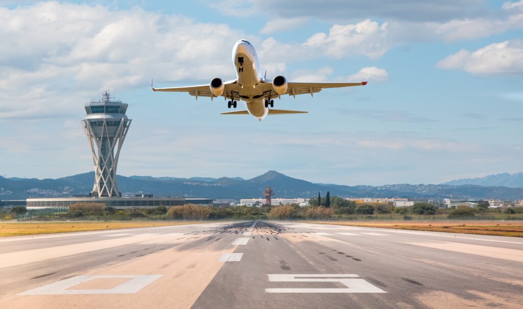 A passenger plane takes off from the runway with the control tower, mountains, and blue sky in the background