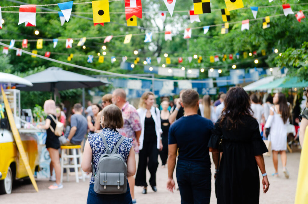 Pedestrians walk through a street festival, with flag pennants hanging overhead and green trees in the background