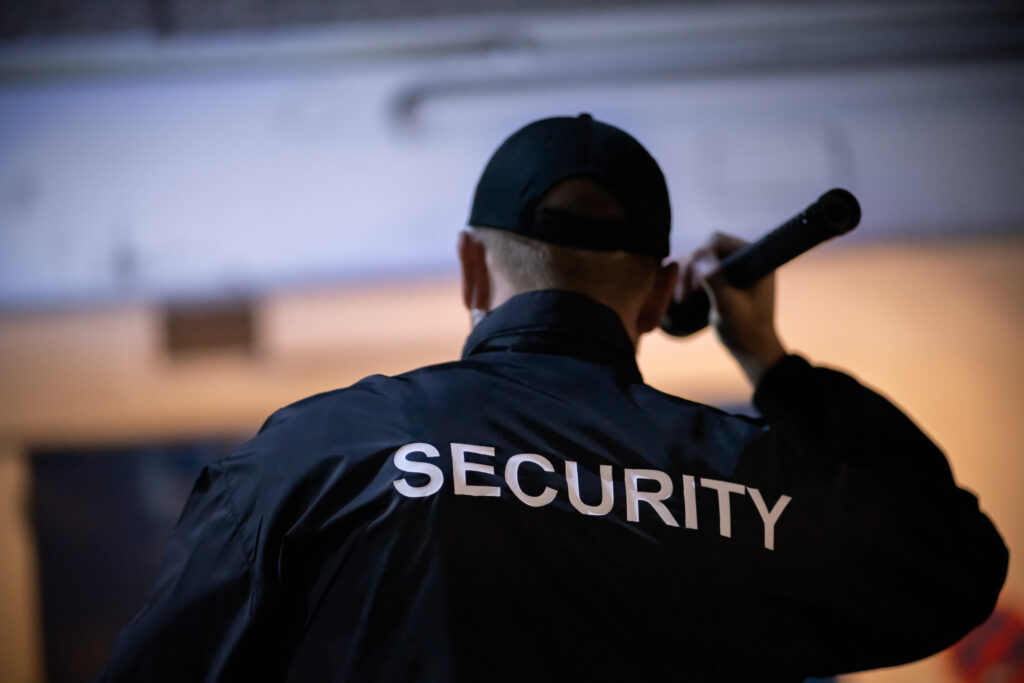 A view of a security guard wearing a dark blue jacket with white lettering holding a flashlight and conducting a security assessment