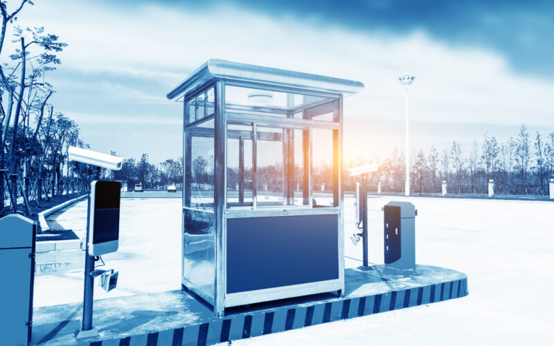 An empty guard booth, surveillance cameras, and access controls at a vehicle security barrier in a parking area