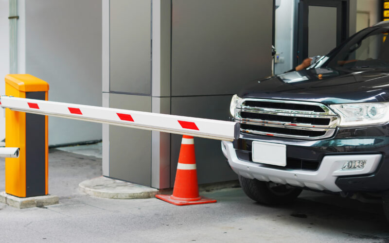 A dark-colored vehicle with a shiny chrome grille waits in front of a crash-rated beam to enter a parking facility.
