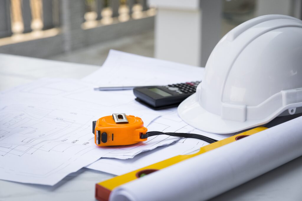 A white hard hat sits on a blueprint with measuring tape illustrating the site security design and planning process.